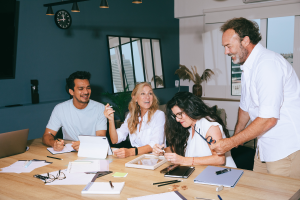 Four people at a table, one of them is standing. All individuals are laughing and having a great time working together on a project. 
